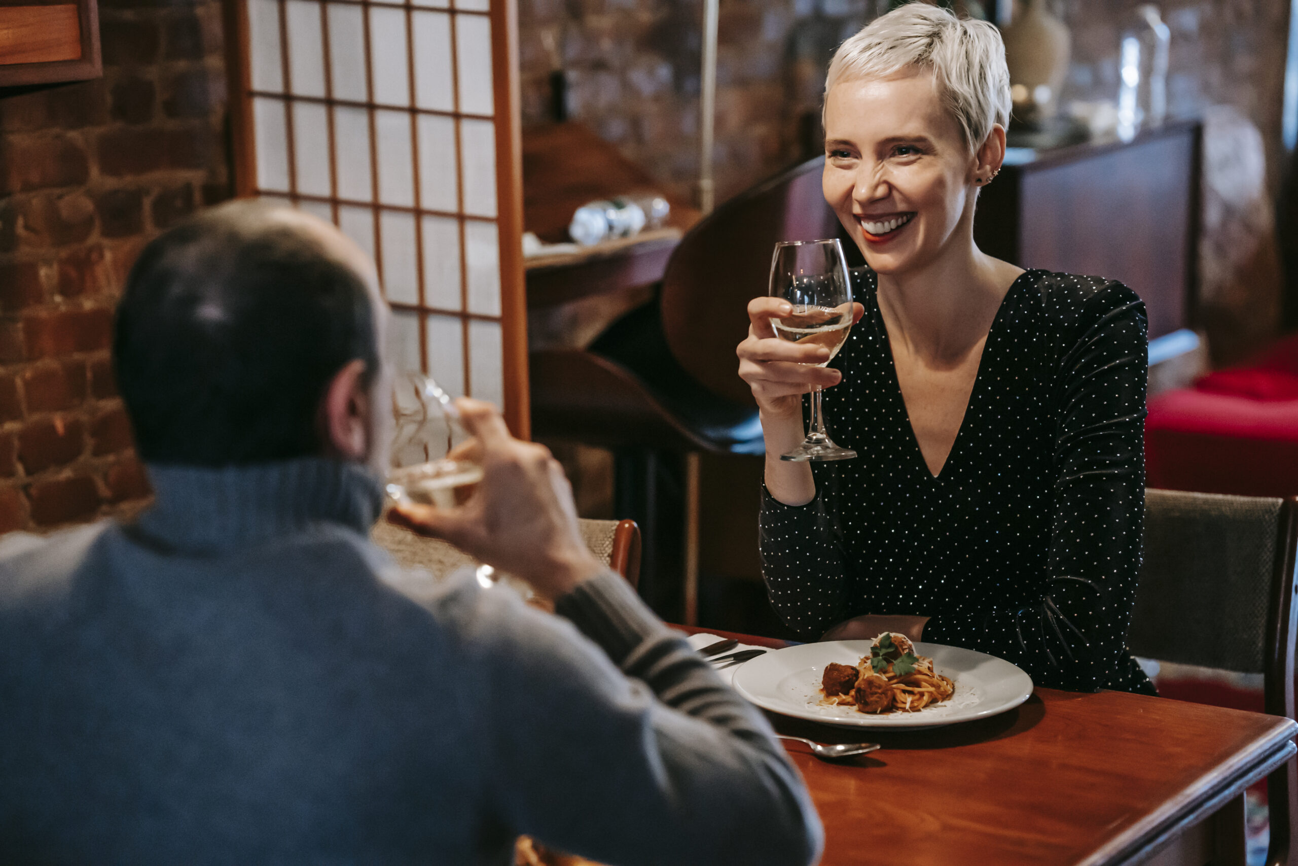 Positive couple sitting in chairs at wooden table while having dinner with pasta with meatballs on plates near cutlery with wineglasses in light room while looking at each other
